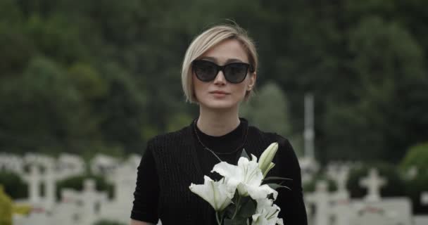 Porträt eines Mädchens mit Sonnenbrille und Trauerkleidung, das in die Kamera blickt. Junge Witwe mit weißer Lilienblüte auf dem Friedhof. Konzept des Gedenktages. Unklarer Hintergrund. — Stockvideo