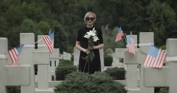 Retrato de mujer sosteniendo una flor de lirio blanco en el cementerio. Viuda en gafas oscuras mirando a la cámara mientras está de pie en la fila de cruces de piedra con banderas americanas. Concepto de día conmemorativo . — Vídeos de Stock