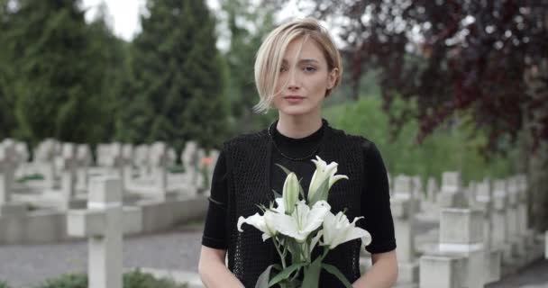 Crop vista della donna in vestiti neri alzando la testa e guardando alla fotocamera. Giovane vedova che regge giglio bianco fiore mentre in piedi al cimitero. Concetto di giorno commemorativo. Sfondo sfocato. — Video Stock