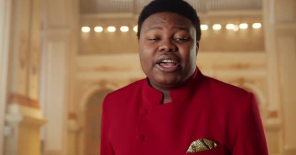 若いアフリカ系アメリカ人男性がゴスペル音楽を歌っている様子を間近で見ることができます。教会に立っている間に感情的に演奏し、手を動かす赤いスーツの男性歌手。人と音楽の概念. — ストック動画
