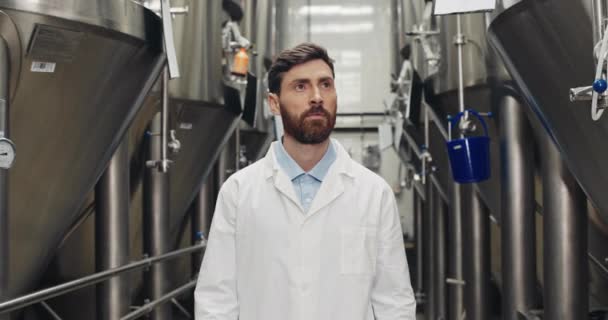 Vista de los cultivos de atento trabajador joven que inspecciona el equipo. Hombre en bata de laboratorio blanca caminando a través de filas de cubas de acero y mirando a un lado. Concepto de fábrica de cerveza y planta de cerveza . — Vídeos de Stock