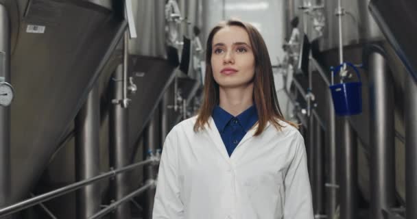Beskär syn på uppmärksam kvinnliga arbetstagare inspektera utrustning. Flicka i vit labbrock går genom rader av stål bryggning kar och tittar åt sidan. Begreppet bryggeri och ölfabrik. — Stockvideo
