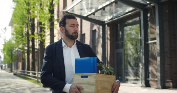Nešťastný muž ve třiceti drží skříňku se svými věcmi, když stojí na ulici. Zaměstnanec ve formálním obleku otočí hlavu a dívá se do kamery. Přiblížit. Pojetí nezaměstnanosti. — Stock video