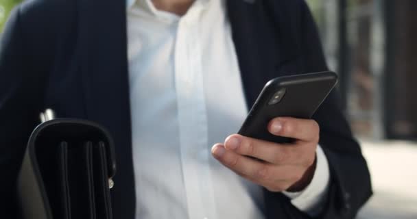 Crop view van mannelijke scrollen telefoon scherm tijdens het surfen op internet. Close-up zicht op de mens in formele kleding met behulp van zijn smartphone tijdens het lopen en draagtas op straat. — Stockvideo