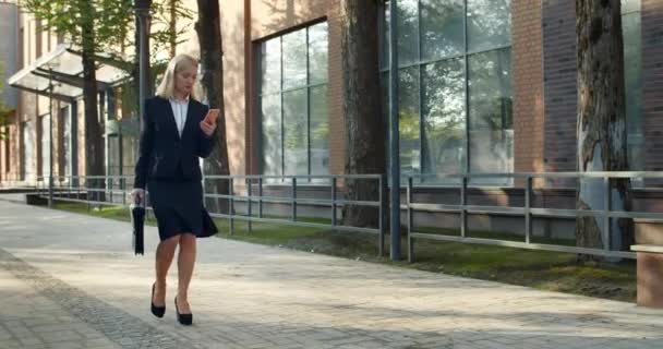 Trabajadora de oficina exitosa en traje formal mirando la pantalla del teléfono mientras camina por la calle del distrito de negocios. Hermosa mujer de negocios que lleva el caso y el uso de su teléfono . — Vídeos de Stock