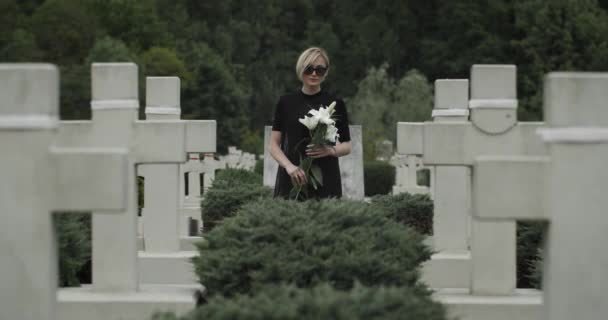Πορτρέτο της νεαρής χήρας κοιτάζοντας στην κάμερα, ενώ στέκεται στη σειρά από πέτρινους σταυρούς. Γυναίκα με σκούρα γυαλιά και ρούχα με λευκά λουλούδια κρίνου στο νεκροταφείο. Έννοια της ημέρας μνήμης. — Αρχείο Βίντεο