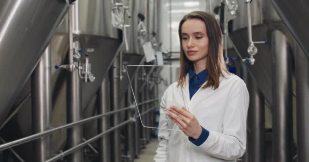 Beskär vy av process engeneer vidröra och titta på tom display. Bryggeri kvinnlig arbetare i labbrock med futuristisk transparent glas tablett när du står på rader av bryggning kar. — Stockvideo