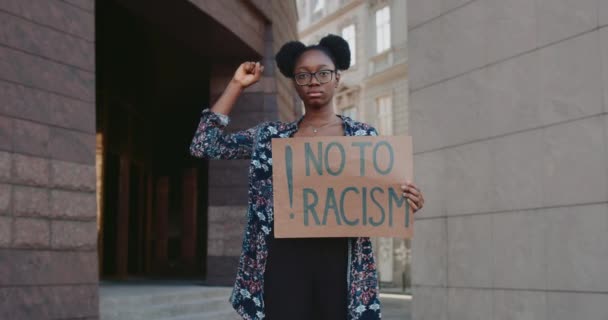 Chica afroamericana alzando el puño apretado y sosteniendo pancarta de cartón sin racismo. Mujer que apoya el movimiento de derechos humanos en pie en la calle de la ciudad. Acercar . — Vídeos de Stock