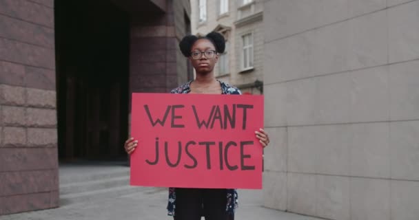 Afro american female student holding placard with we want justice writing on it. Serious girl in glasses supporting human rights movement while standing at city street. Zoom in. — Stock Video