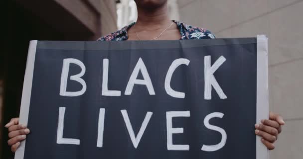 Vista de cerca de la niña afroamericana sosteniendo pancarta con la escritura de la materia de vidas negras. Activista femenina que apoya el movimiento contra la brutalidad y el racismo estando de pie en la calle . — Vídeo de stock