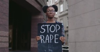Afro-Amerikan kız portresi. Elinde karton kutu var. Şehir caddesinde tecavüz sloganı var. Şiddete ve saldırıya karşı hareketi destekleyen kadın eylemci. Sosyal sorunlar kavramı.