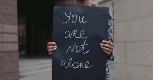 Female african american hands holding carton placard with you are not alone writing on it at city street. Concept of social problem and support. — Stock Video