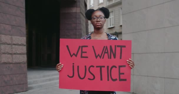 Portret van een vrouwelijke activiste die een spandoek vasthoudt met gerechtigheid erop. Afro-Amerikaans meisje in bril ter ondersteuning van de mensenrechten beweging terwijl staan op lege stad straat. — Stockvideo