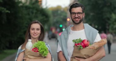 Sokakta yürüyen ve ellerinde yiyecek dolu kağıt torbalarla gezen neşeli çiftin mahsul manzarası. Güzel bir adam ve kadın kameraya bakar ve alışveriş yaparken gülümserler..