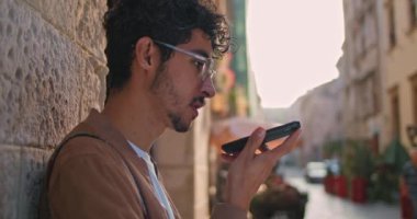 Gözlüklü bir gencin duvara yaslanmış bir şekilde hoparlörde yazarken ve konuşurken görüntüsünü kapat. Milenyum çocuğu internette geziniyor ve caddede akıllı telefon ses asistanı kullanıyor..