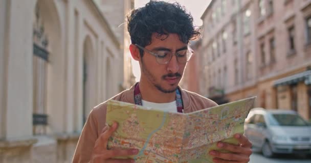 一个戴着眼镜的年轻人一边走路一边看一边看的作物图片。一个英俊的家伙带着鼻环搜索地址和地图在老城区街道上。旅行和旅游的概念. — 图库视频影像