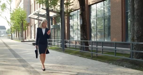 Good looking female employee in formal clotes looking at phone screen while scrolling news feeds. Businesswoman in 30s using her mobilephone while walking and carrying case at street. — Stock Video