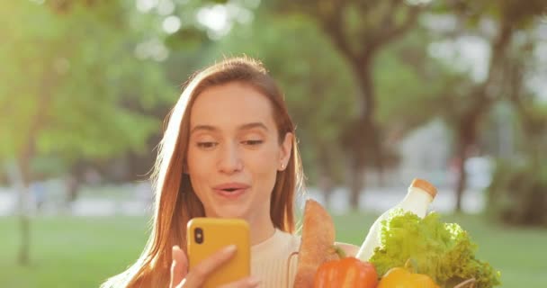 Vista de cerca de la hermosa mujer joven caminando y leyendo buenas noticias en el teléfono inteligente con la cara sorprendida. Chica sorprendida llevando comida en una bolsa de papel y mirando la pantalla del teléfono. Al aire libre . — Vídeo de stock