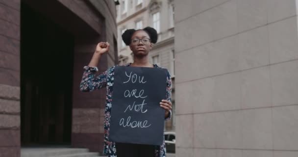 Női aktivista kezében placard veled nem egyedül írni, miközben a kamerába néz. Az afro-amerikai lány ökölbe szorított öklöt emel, miközben az utcán őrködik. A társadalmi probléma fogalma. — Stock videók