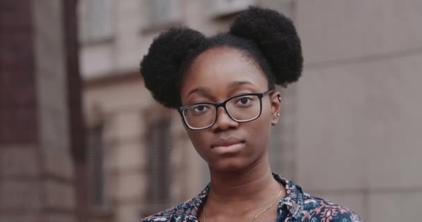 Vista da vicino della ragazza afro-americana in occhiali che guarda alla fotocamera con una seria espressione facciale. Ritratto di giovane ragazza in piedi sulla strada della città.Concetto di persone ed emozioni. — Video Stock