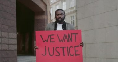 Afro-Amerikan erkek eylemci pankart tutuyor. Üzerinde adalet yazısı olsun istiyoruz. Şehir caddesinde dururken insan hakları hareketini destekleyen ciddi sakallı adam. Yakınlaştır.