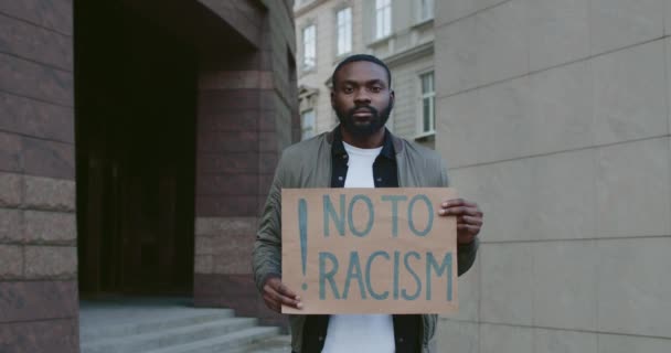 Retrato del chico afroamericano mirando a la cámara mientras sostiene la pancarta no al racismo. Hombre barbudo protestando contra la desigualdad racial en la calle de la ciudad. Concepto de derechos humanos . — Vídeo de stock