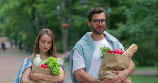 Vista frontal de pareja joven mirando a la cámara mientras lleva bolsas de papel con comida y sonriendo. Tipo barbudo en gafas y su encantadora novia caminando en el parque después de ir de compras. Al aire libre . — Vídeos de Stock