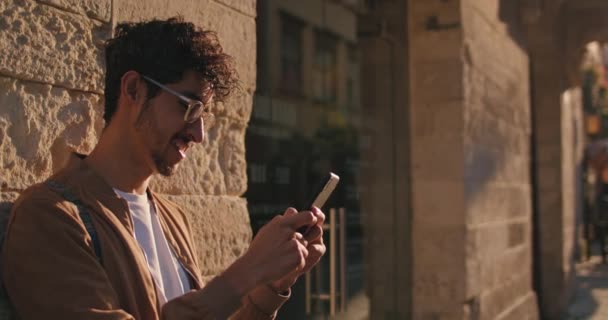 Crop view van vrolijke man in glazen met behulp van smartphone terwijl staande leunde aan de muur. Jongeman met neusring typen en glimlachen tijdens het chatten in sociale netwerken in de stad straat. — Stockvideo