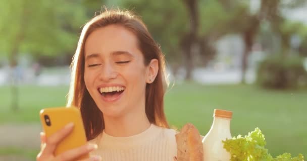 Mulher millennial alegre rindo enquanto olha para a tela do smartphone e lê uma mensagem engraçada. Close-up vista da bela menina sorridente carregando comida enquanto caminha e usando seu telefone . — Vídeo de Stock