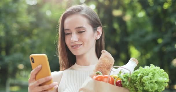 Millennial aantrekkelijke meisje opknoping in sociale netwerken terwijl staan in de straat en het houden van papieren zak met boodschappen. Vrolijke jonge vrouwen scrollen nieuws feed tijdens het gebruik van de smartphone. — Stockvideo