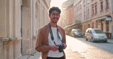 Elinde kamera olan ve gülümseyen bin yıllık erkek turistin portresi. Eski şehir sokağında güneş ışınlarıyla durup poz veren gözlüklü yakışıklı bir adam. Seyahat ve turizm kavramı.