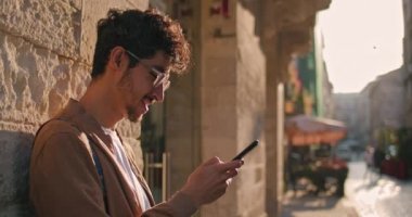 Gözlüklü yakışıklı bir erkek turist duvara yaslanırken cep telefonu kullanıyor. Şehir caddesinde kaydırma ve daktilo ekranı kullanırken gülümseyen bin yıllık bir adamın ekili görüntüsü.