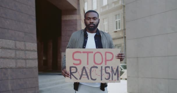 Portrait of afro american bearded guy holding stop racism phrase cardboard. Handsome male activist supporting anti racism campaign at city street. Concept of equal human rights. — Stock Video