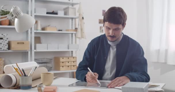テーブルの上に描かれた口ひげを生やした男。居心地の良い職場でテーブルに座っている間、デジタルノートパッドとスタイラスを使用して若い男イラストレーター。創造性の概念. — ストック動画