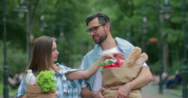 Egy boldog pár, akik visszajönnek a boltból, és papírzacskókat tartanak tele kajával. Csinos nő gránátalmával és hányással sétál a férjével. Szabadtéri. — Stock videók