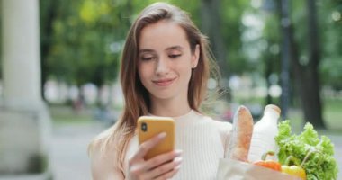 Bin yıllık güzel bir kadının alışveriş torbalarıyla kağıt torba taşırken internete göz atmasını yakından izleyin. Akıllı telefon kullanan ve dışarıda dolaşan çekici bir genç kız. Teknoloji ve alışveriş kavramı.