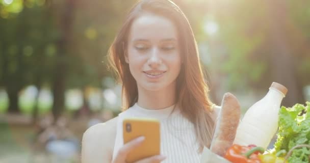 Närbild bild av glad flicka surfa nätet och ler medan du använder sin smartphone.Charming ung kvinna bär sina livsmedel i papperspåse när du går och tittar på skärmen. Utomhus. — Stockvideo