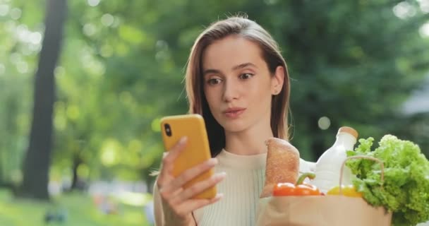 Gros plan de brunnette fille obtenir de mauvaises nouvelles sur son smartphone et l'air choqué. Fille bouleversée tenant sac en papier avec épicerie et regardant l'écran du téléphone tout en se tenant debout à la rue. — Video