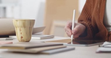 Grafik tabletindeki kadın web tasarımcısının görüntüsünü kapat, masada otururken ekrana dokun ve yakınlaştır. Stylus kullanan ve işyerindeki bardaktan içen insanlar..