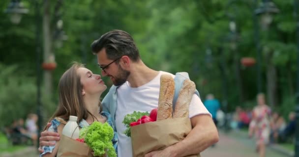 Γλυκό ζευγάρι κουβαλάει χάρτινες σακούλες με φαγητό και περνάει καλά μαζί. Όμορφος γενειοφόρος άντρας με γυαλιά αγκαλιάζει και φιλάει την ελκυστική γυναίκα του ενώ περπατούν στο πάρκο.. — Αρχείο Βίντεο