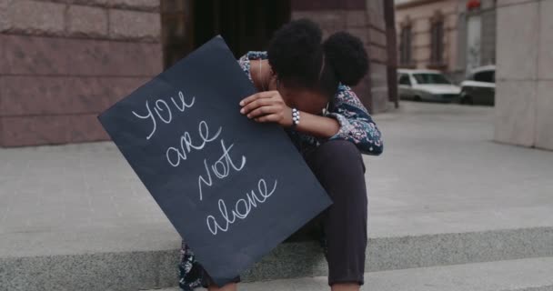 Chica afroamericana deprimida poniendo la cabeza de rodillas mientras está sentada en los escalones. Mujer joven sosteniendo cartel de cartón con usted no está escribiendo solo. Concepto de problema social . — Vídeos de Stock