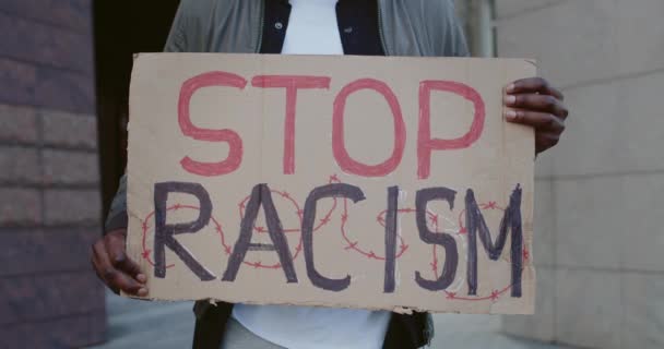 Vista de las cosechas de las manos afroamericanas que sostienen pancarta de cartón con escritura de alto el racismo. Activista masculino protestando en la calle de la ciudad. Concepto de derechos humanos y desigualdad racial . — Vídeos de Stock