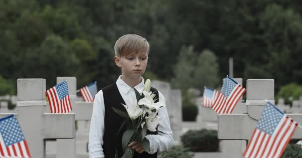 Zblízka dospívající chlapec drží lilie květiny, zatímco stojí u hrobu svého otce. Dítě uctívající svého otce vojáka na hřbitově. Koncept vzpomínkového dne. Americké dlaždice u kamenných křížů. — Stock video