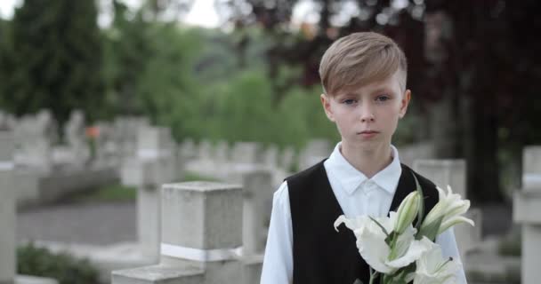 Портрет засмученого хлопчика, який піднімає голову і дивиться на камеру. Вид на урожай молодої дитини, що тримає білу квітку лілії, стоячи на цвинтарі. Концепція пам'ятного дня . — стокове відео