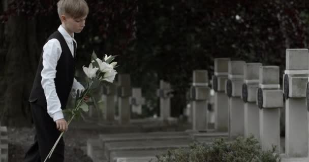 Seitenansicht eines kleinen Jungen, der weiße Lilienblumen auf den Grabstein seines Vaters legt. Teenager-Kind zu Ehren seines Vaters Soldaten auf dem Friedhof. Konzept des Volkstrauertages. — Stockvideo