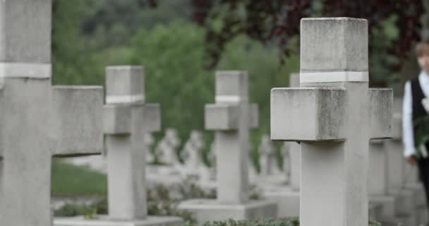 Tonårspojken håller vita liljeblommor när han går på kyrkogården. Begreppet minnesdag. Gravstenar på bakgrunden. — Stockvideo