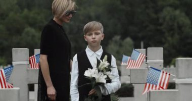Koyu renk elbiseli bir anne mezarlıkta dikilen oğluna sarılıyor. Elinde çiçekler olan genç bir çocuk Amerikan bayraklarıyla sıralanmış taş haçlara bakıyor. Anma Günü kavramı.