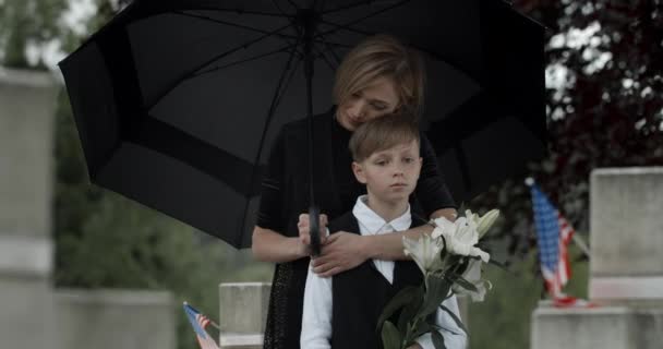 Θλιβερή γυναίκα αγκαλιάζει και γέρνει προς το έφηβο παιδί του ενώ στέκεται κάτω από ομπρέλα στο νεκροταφείο.Χήρα και νεαρό αγόρι με λευκά λουλούδια κοντά σε ταφόπλακα με αμερικανική σημαία.Έννοια της ημέρας μνήμης. — Αρχείο Βίντεο
