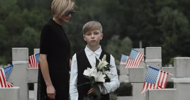 Moeder in donkere kleren komen en knuffelen zoon staan op begraafplaats.Tiener jongen met bloemen en kijken naar camera in rij van stenen kruisen met Amerikaanse vlaggen. Begrip herdenkingsdag. — Stockvideo