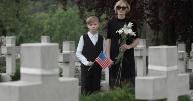 Amerikan bayrağı tutan çocuğun portresi ve kameraya bakan çiçekli genç bir kadın. Mezarlıkta dikilirken oğlunun elini tutan koyu renk elbiseli bir kadın. Anma Günü kavramı.